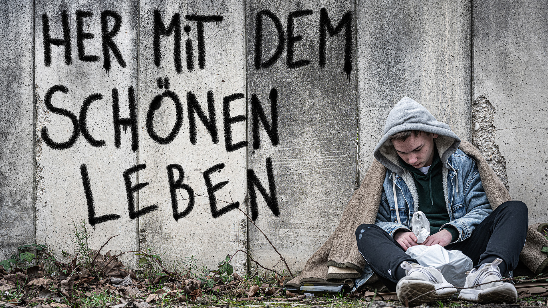 Ein Jugendlicher sitzt auf dem Boden vor einer Mauer mit der Aufschrift „Her mit dem schönen Leben“