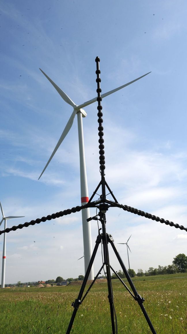 Eine dreiarmige akustische Kamera steht in der Nähe von Windkrafträdern auf einem Feld.