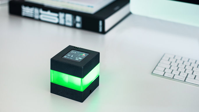Ein würfelförmiges Gerät, das farbig leuchtet, auf einem Schreibtisch neben einer Tastatur und einem Ordner