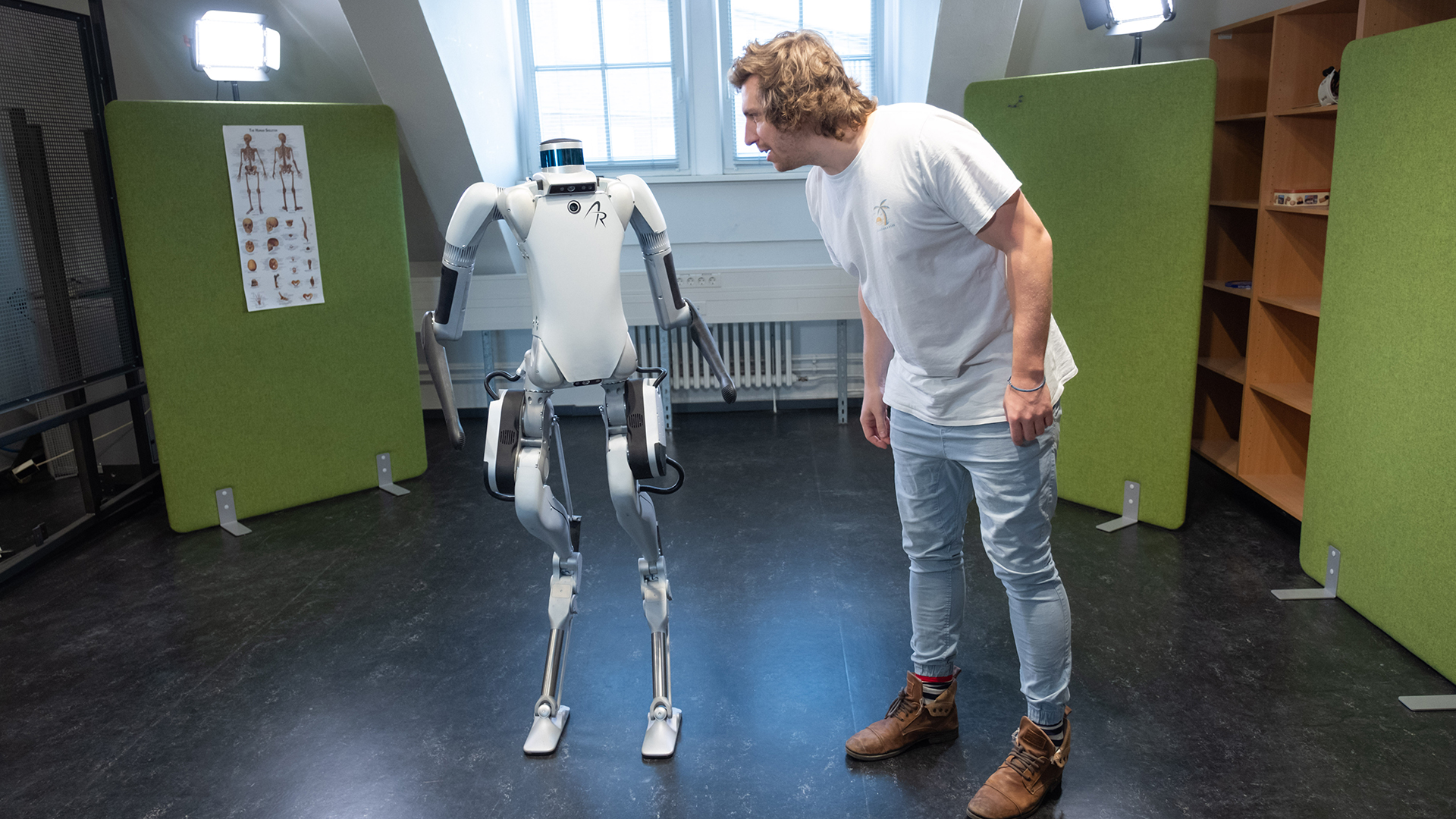 In einem weitgehend leeren Raum stehen ein zweibeiniger Roboter und ein Mensch nebeneinander und scheinen miteinander zu sprechen.