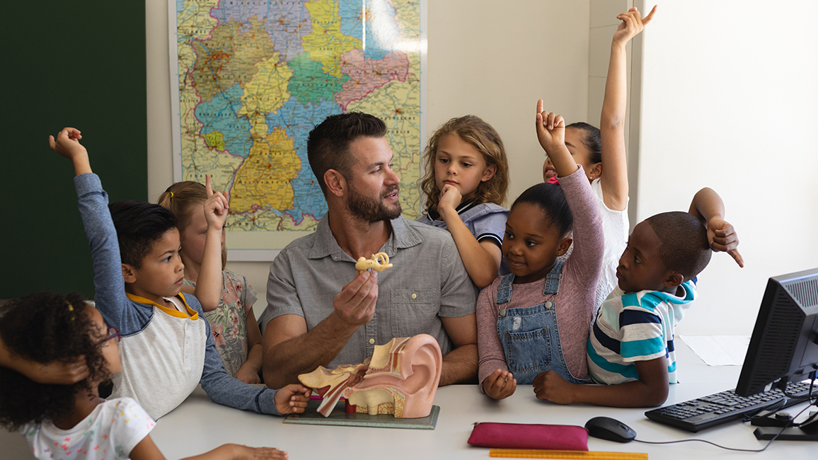 Ein Lehrer sitzt an einem Tisch, auf dem ein Modell von einem menschlichen Ohr steht; um ihn herum Grundschulkinder, die aufzeigen oder zuhören.