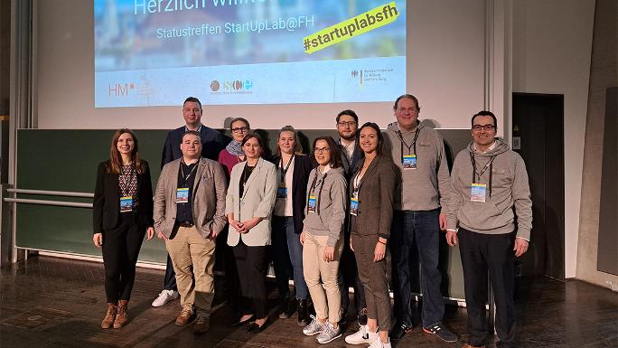 Elf Personen stehen auf einer Bühne, über ihnen auf der Leinwand eine Präsentation mit dem Schriftzug "Statustreffen StartUpLab@FH" und dem Hashtag #startuplabsfh.