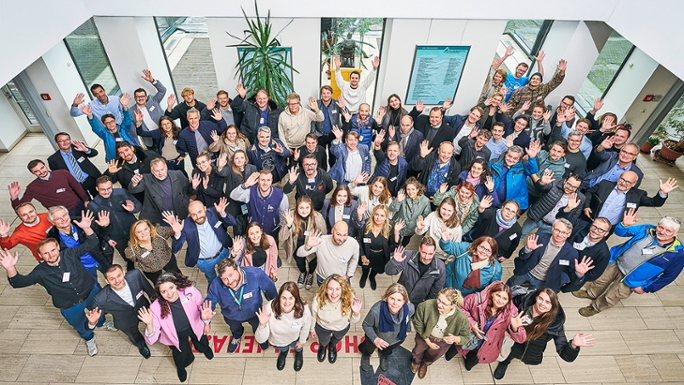 Eine Gruppe von etwa 50 Personen steht winkend in einem Foyer und wird dabei von schräg oben fotografiert