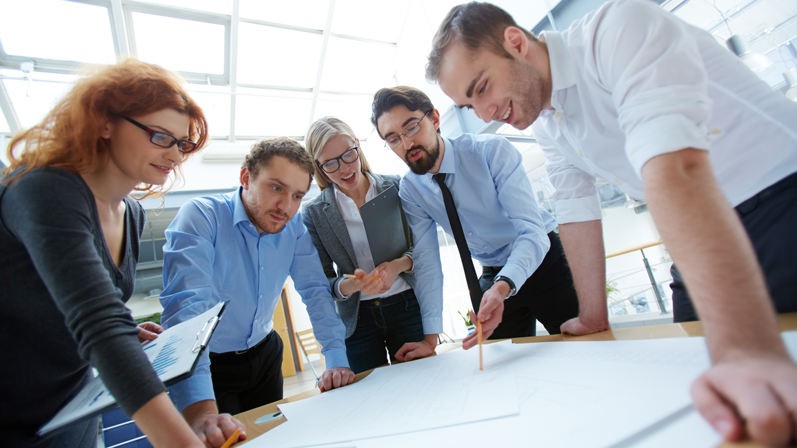 Five men and women stand bent over a table in a semicircle and discuss a concept