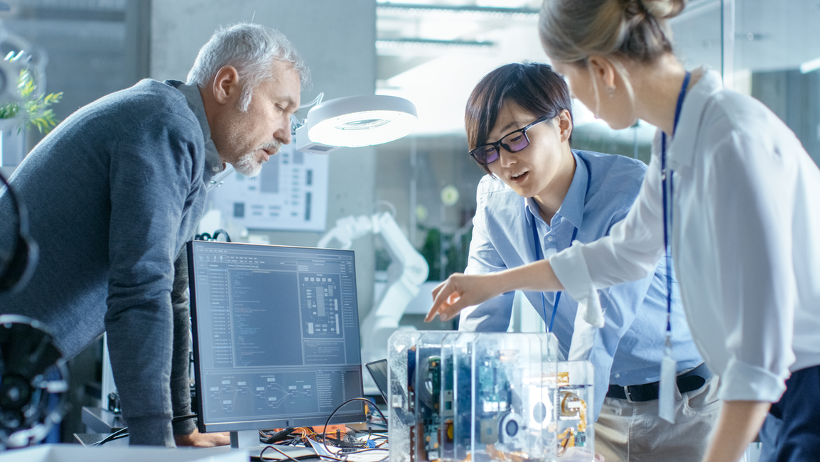 Three people stand at a table full of technology and computers, one person explains something to the others. Atmosphere: technical