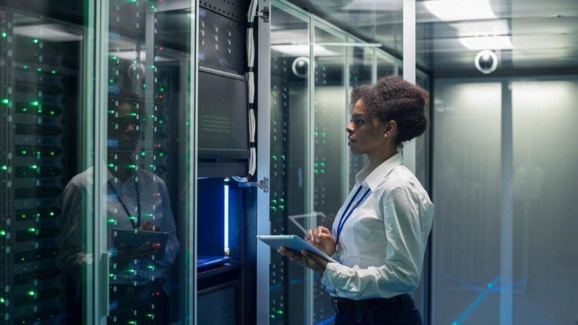 A young woman stands in front of a server cabinet with a tablet in her hand