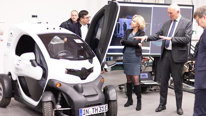 On the left of the picture is a small car. To the right, a group of people are looking at the car. There is a large screen in the background.