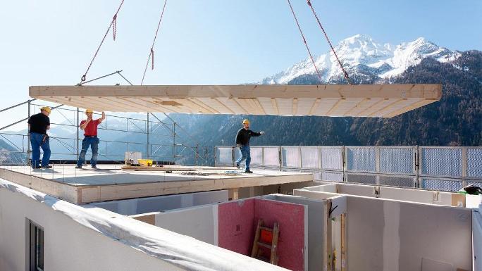 A ceiling element of a prefabricated house hovers on a crane above a building shell and is directed to the right position by three construction workers.
