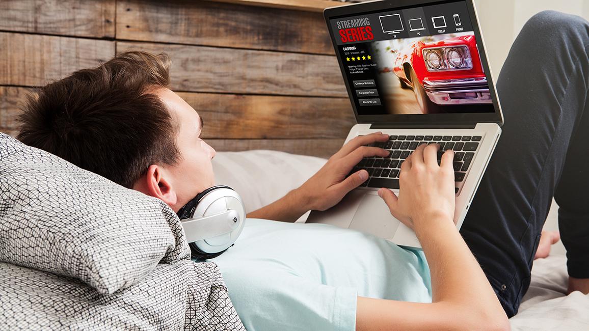 A man lies on a sofa with a laptop on his lap. The screen shows a streaming website.