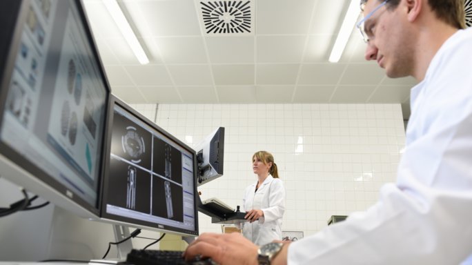 A man in a lab coat sits at a double monitor, photographed from below