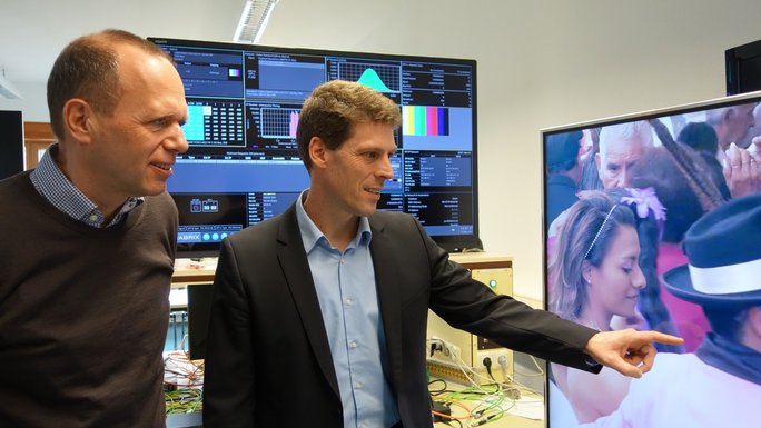 Two men stand in front of a big screen where a series is streaming. In the background there are technical devices like a computer and another big screen which displays technical data.